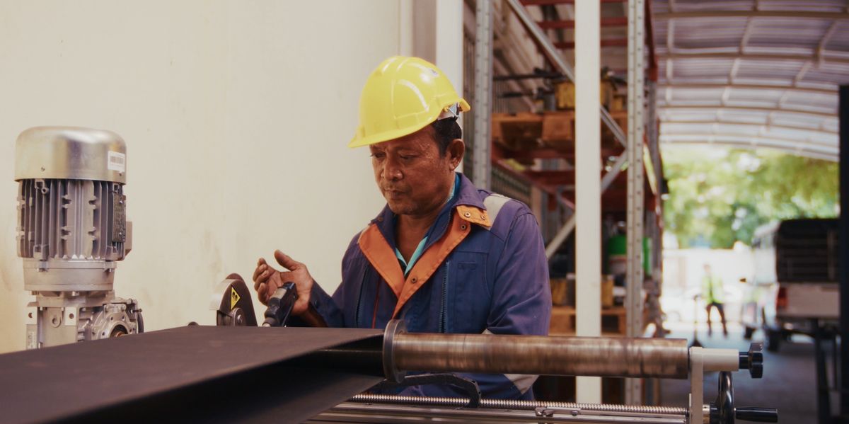 Worker in a yellow hard hat and blue coveralls inspects a metal pipe on a fabrication line inside a workshop