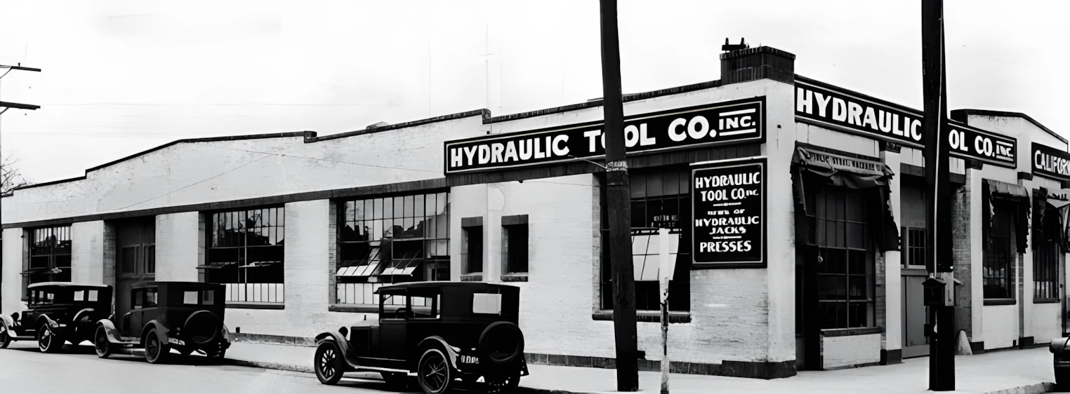Historic street scene featuring a white brick hydraulic tools building with vintage cars parked along the curb (black-and-white photo).