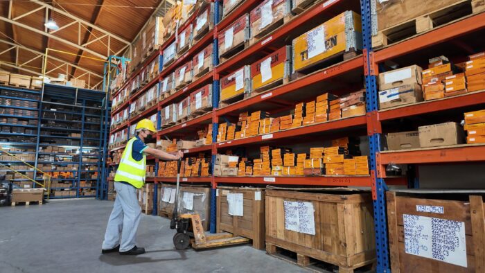 Warehouse worker in a yellow hard hat and reflective vest using a pallet jack beside tall orange shelving filled with labeled boxes in a warehouse
