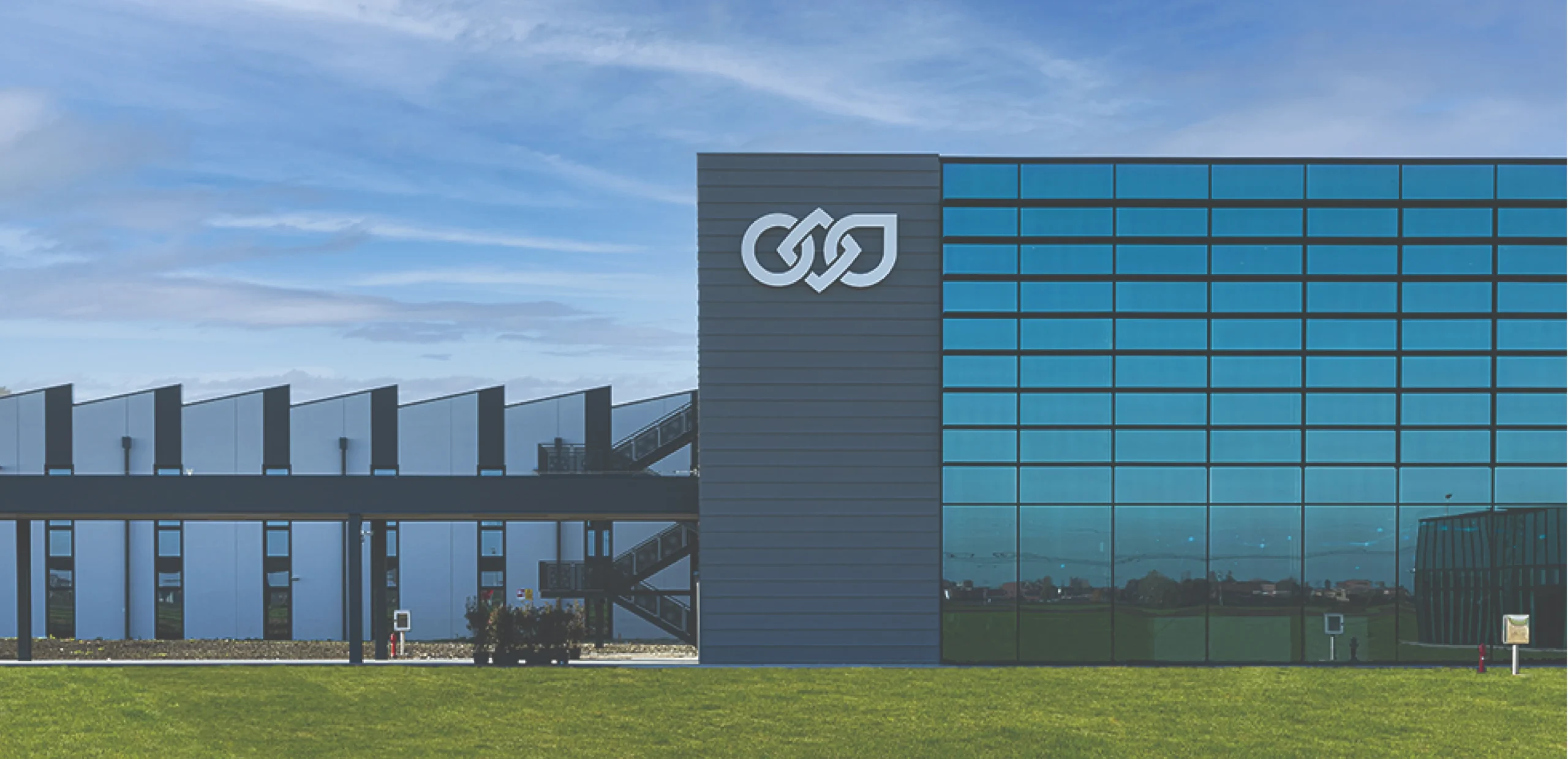 Modern office building with blue glass windows and a white intertwined logo on a gray panel, grass in the foreground.