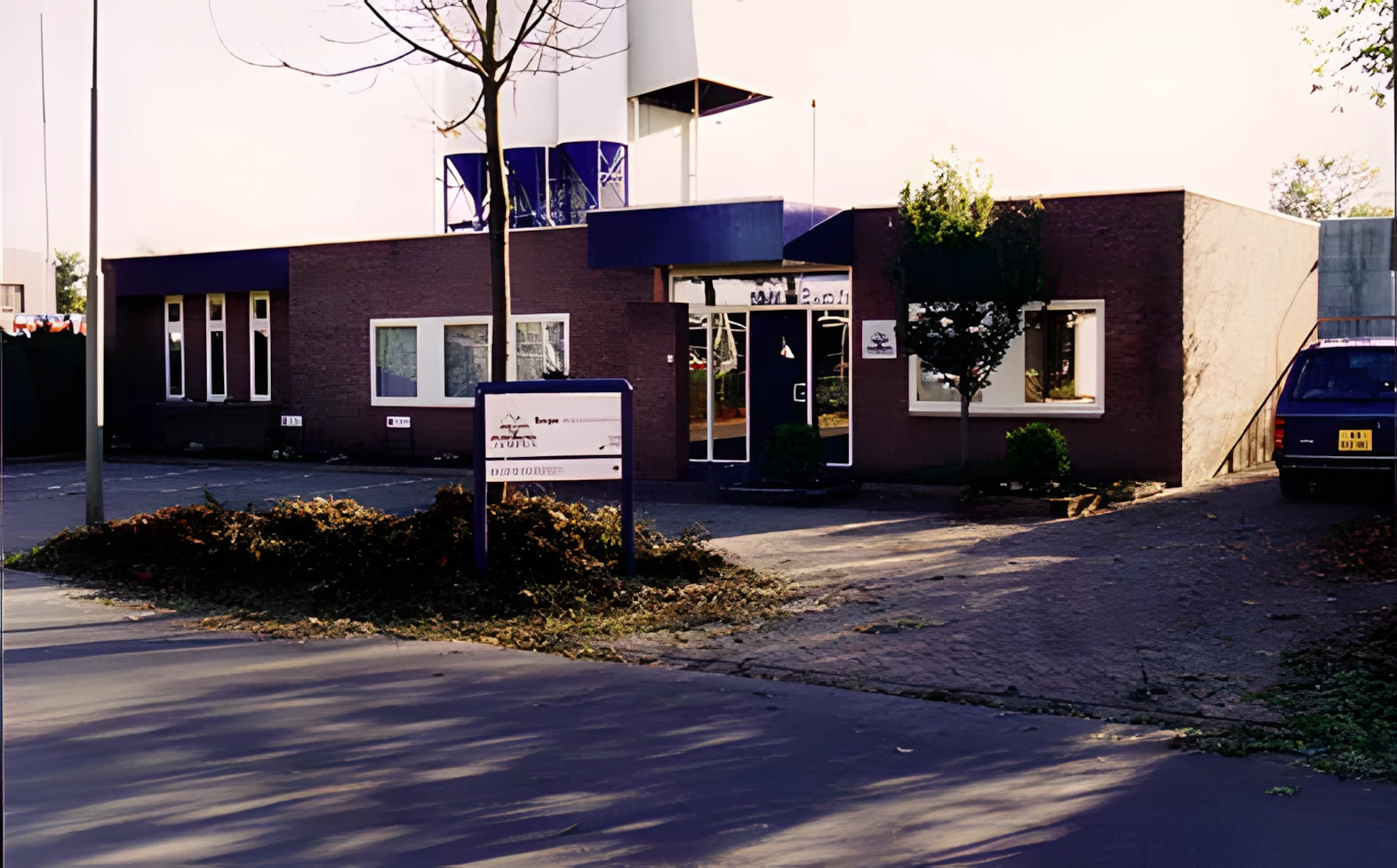 Brick, single-story building with a glass entrance and blue awning; a front sign, and a blue SUV parked to the right with a tree near the door.