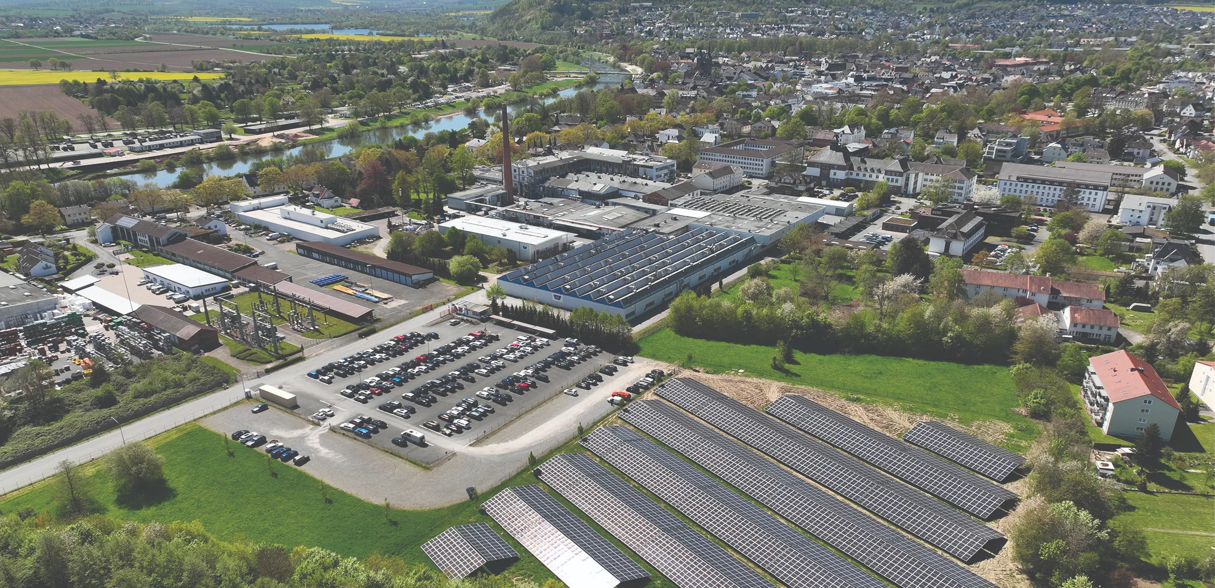 Aerial view of a town with a winding river, industrial buildings with a tall smokestack, and a large solar panel field in the foreground.